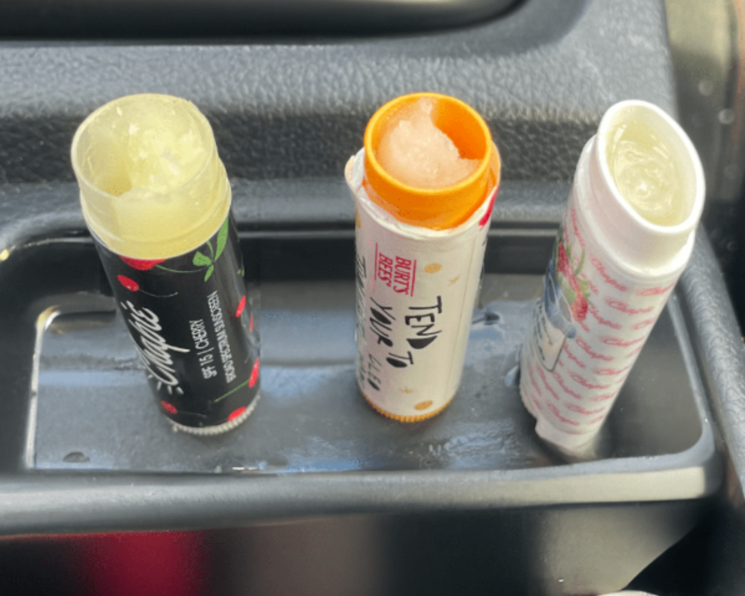 Three open lip balm tubes placed in the cup holder of a car interior. The left tube is black with red fruit graphics and reads 'Watermelon Lip Balm'; the middle tube is white with an orange lid and reads 'TED BEE'; the right tube is white with pink graphics.