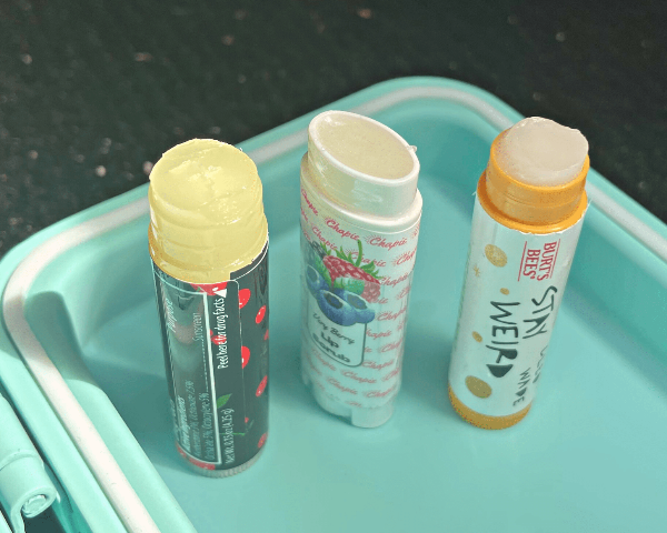Three lip balm tubes standing on a mint green plastic tray. The left tube has a yellow balm and a dark red/black label with red strawberry illustrations.